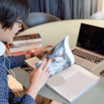 A student pores over a textbook with their computer open as they write their cover letter